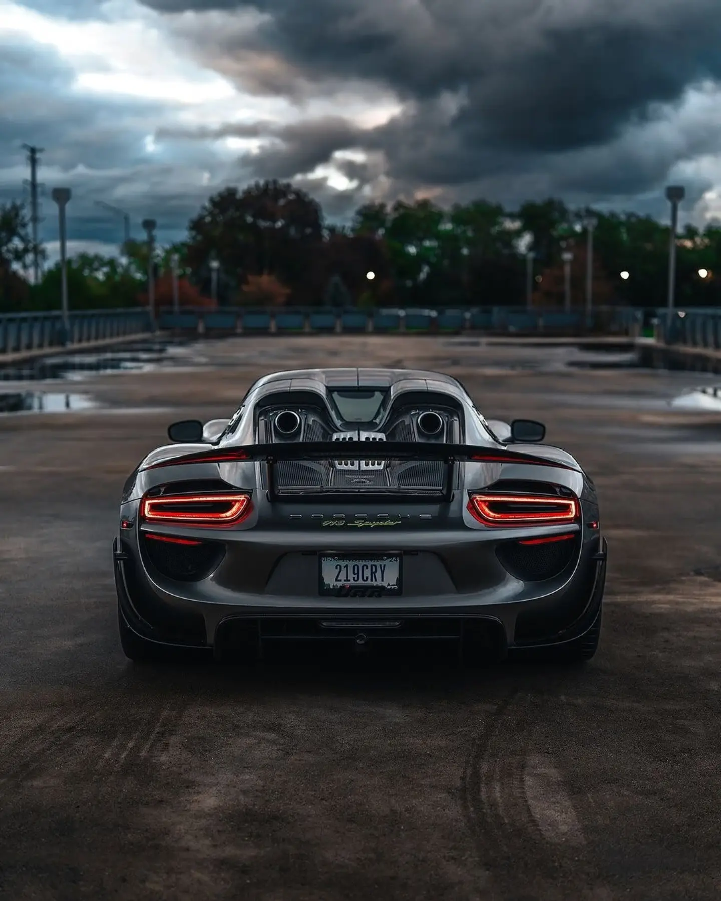 Heckansicht eines grauen Porsche 918 Spyder auf einem nassen Parkdeck unter dunklen Wolken