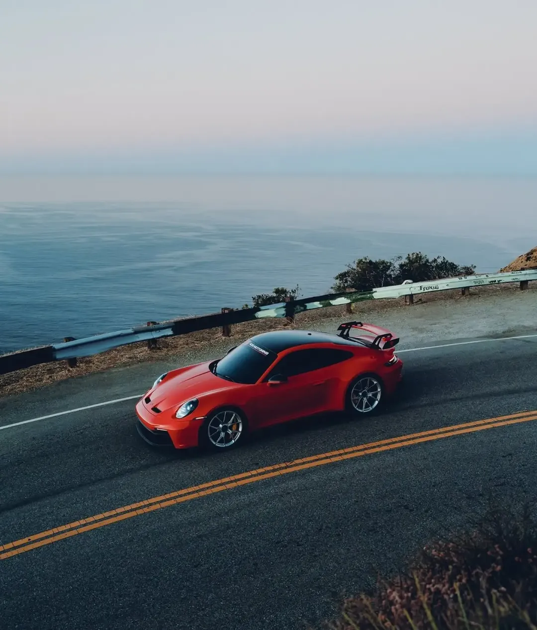 Roter Porsche mit Heckflügel auf einer Küstenstraße am Meer aus erhöhter Perspektive