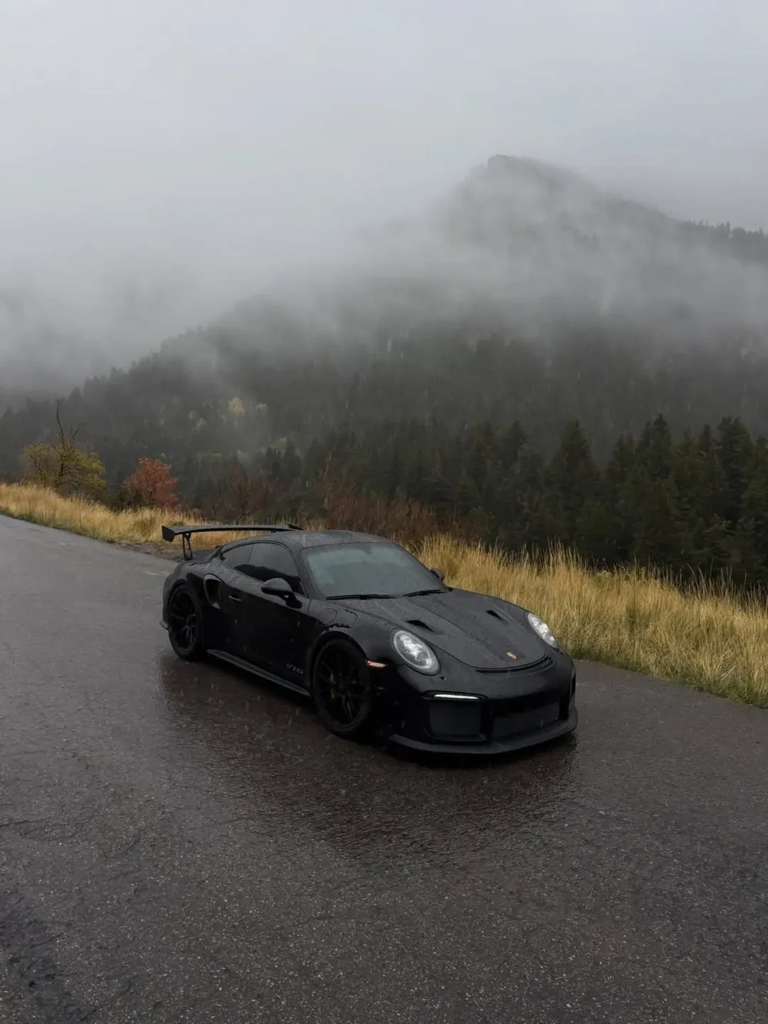 Schwarzer Porsche mit großem Heckflügel auf einer nassen Bergstraße vor nebligen Waldhängen