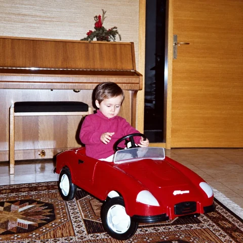 Als KI-Inhalt gekennzeichnetes Rückblickmotiv: Kind in einem roten Porsche-Tretauto vor einem Klavier