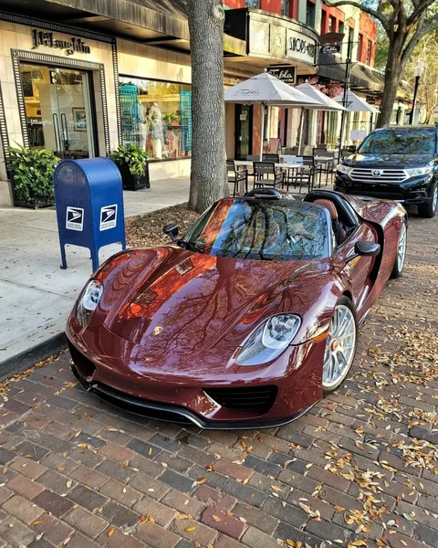 Offener burgunderroter Porsche 918 Spyder an einer Stadtstraße vor Geschäften und Bäumen