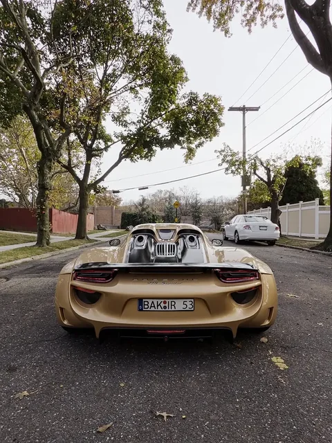 Designmotiv eines goldfarbenen Porsche 918 Spyder in Heckansicht auf einer von Bäumen gesäumten Straße