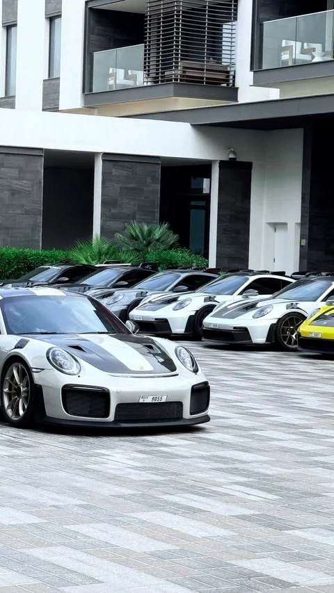 Mehrere Porsche-911-GT-Modelle vor einem modernen Gebäude; im Vordergrund ein weißer Sportwagen mit dunkler Motorhaube. Original-Reelvorschaubild von @speedframe_stories.