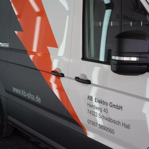 Es ist eine Flottenbeschriftung auf einem VW Crafter in grau und orange zu sehen. Man sieht, das es Folie ist, da man es so nie lackieren könnte. Kleine Schriften auf dem Fahrzeug zeigen, dass es von der Firma KB Elektro GmbH in Schwäbisch Hall ist.