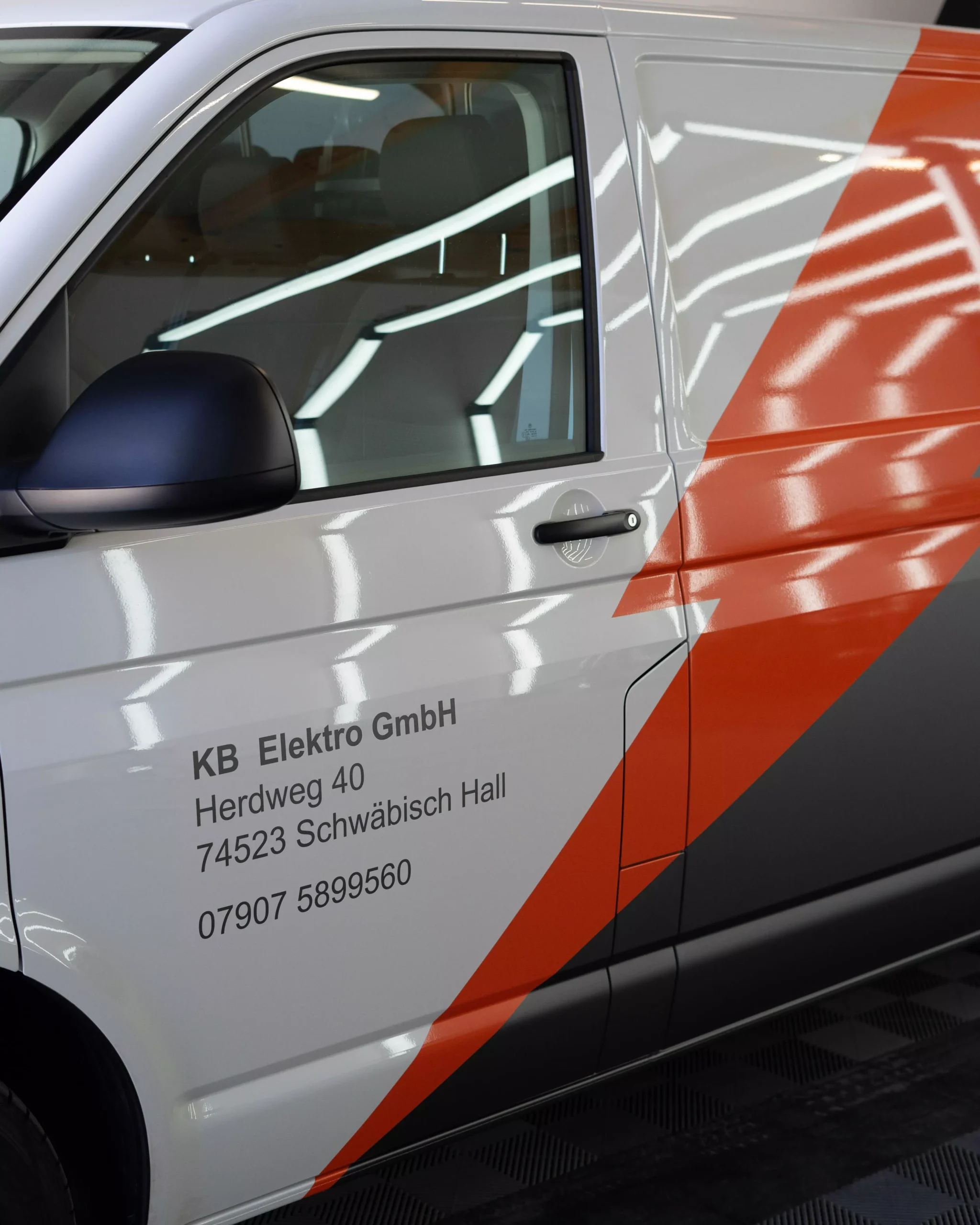 Es ist eine Flottenbeschriftung auf einem VW T6 in grau und orange zu sehen. Man sieht, das es Folie ist, da man es so nie lackieren könnte. Kleine Schriften auf dem Fahrzeug zeigen, dass es von der Firma KB Elektro GmbH in Schwäbisch Hall ist.
