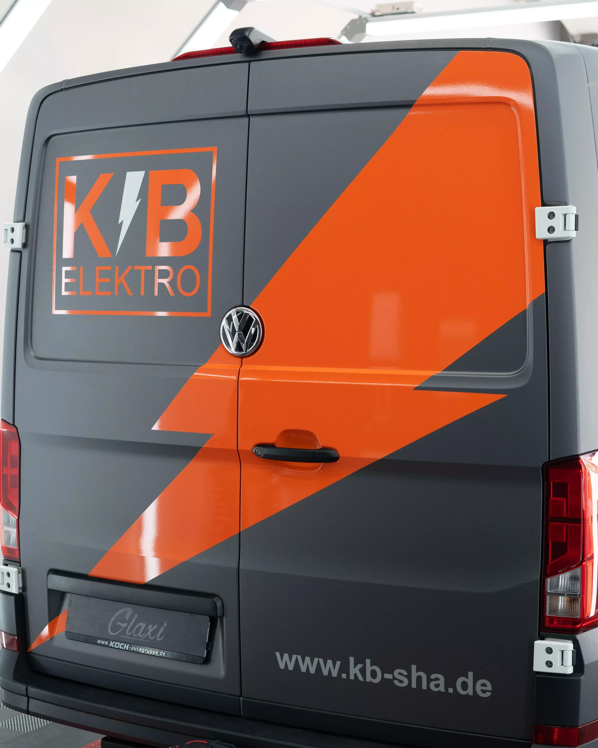 Es ist eine Flottenbeschriftung auf einem VW Crafter in grau und orange zu sehen. Man sieht, das es Folie ist, da man es so nie lackieren könnte. Kleine Schriften auf dem Fahrzeug zeigen, dass es von der Firma KB Elektro GmbH in Schwäbisch Hall ist.