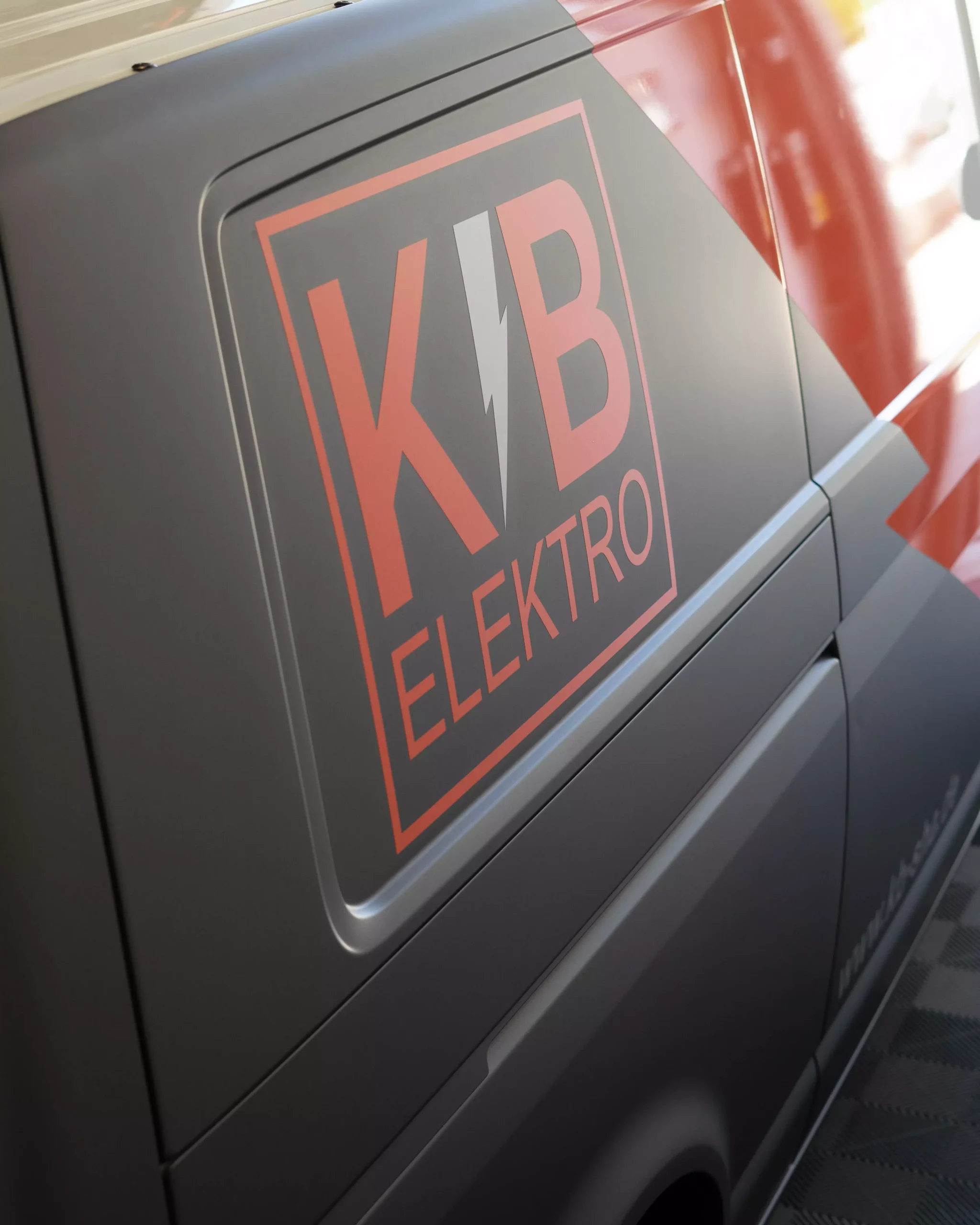 Es ist eine Flottenbeschriftung auf einem VW T6 in grau und orange zu sehen. Man sieht, das es Folie ist, da man es so nie lackieren könnte. Kleine Schriften auf dem Fahrzeug zeigen, dass es von der Firma KB Elektro GmbH in Schwäbisch Hall ist.